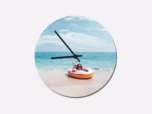 Die runde Wanduhr mit Foto zeigt ein Kind im Schwimmring am Strand vor dem Meer und kombiniert Uhrzeit mit Urlaubsstimmung.
