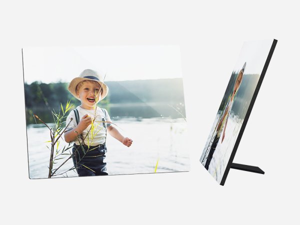 Ein quadratischer Fotoaufsteller zeigt ein fröhliches kleines Kind, das am Ufer eines Sees oder Flusses steht. Das Kind hat blonde Haare und trägt einen hellen Hut, ein weißes T-Shirt mit einem bunten Aufdruck und dunkle Latzhosen. 